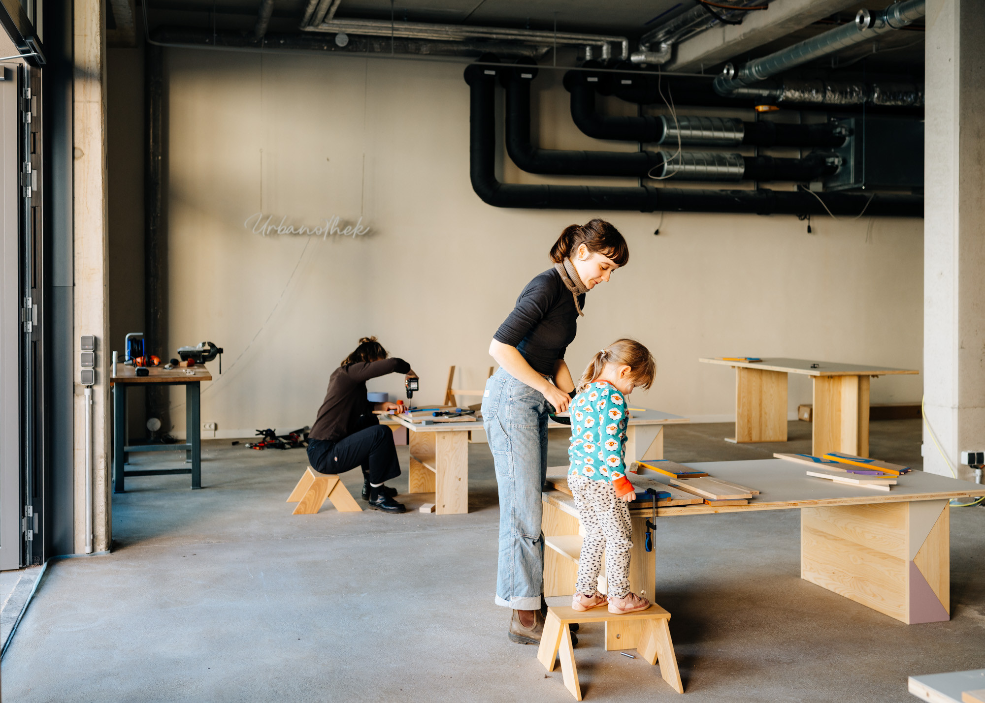 Ein Raum in dem Kindertische und Hocker stehen. Zwei Frauen und ein M&auml;dchen arbeiten mit Holz. Im Hintergrund der Schriftzug Urbanothek.