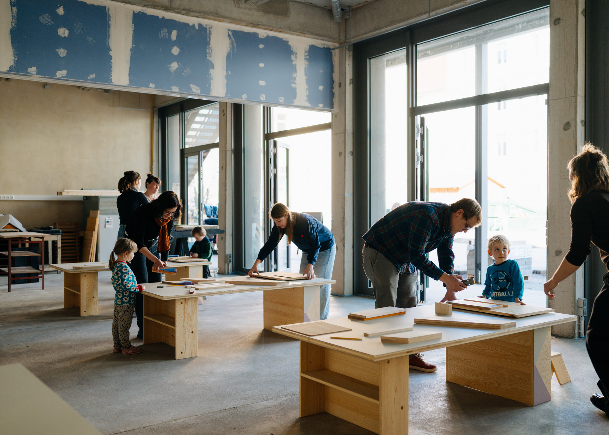 Ein gro&szlig;er Werkstattraum mit Kindertischen aus Holz, in dem ein Workshop mit mehreren Kindern und Erwachsenen stattfindet.