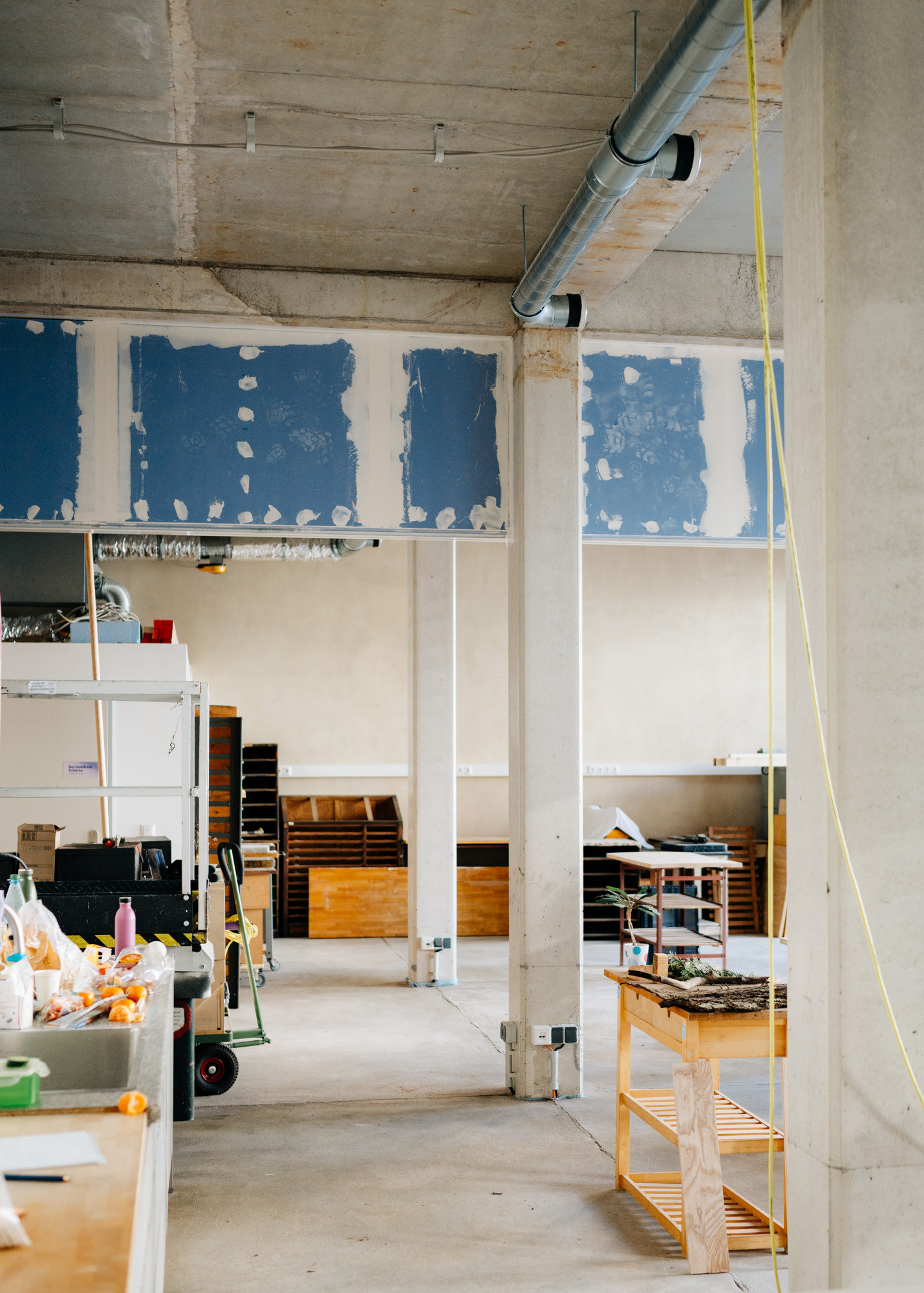 Ein hoher Werkstattraum mit Betonestrichboden S&auml;ulen, Holzm&ouml;beln und einer bunten K&uuml;che.