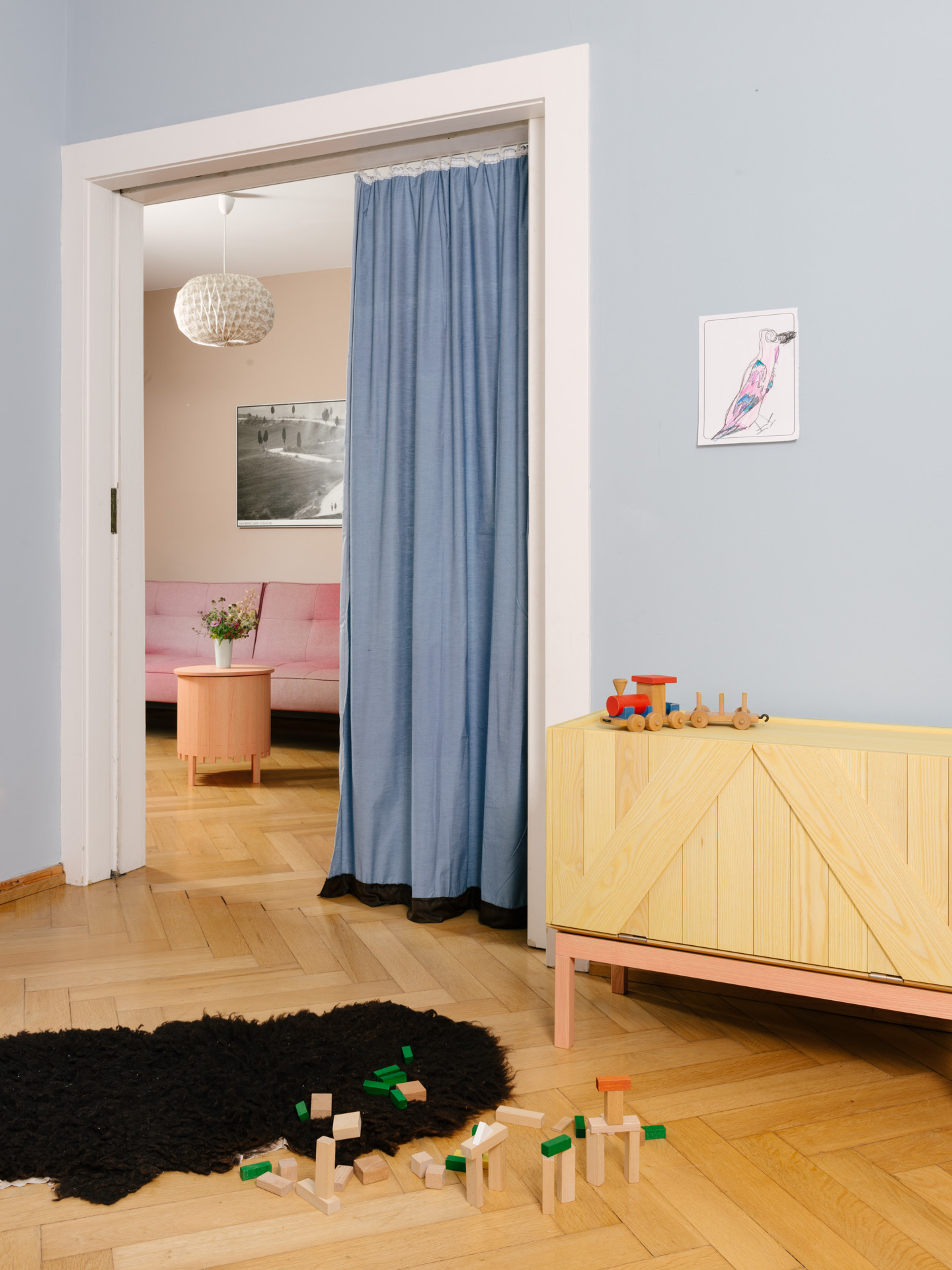 Ein offenes Altbau Kinderzimmer mit erdbeerrosa und zitronengelb lasierten Holzmöbeln (Ein Tischchen und eine Kommode) vor hellblauer Wand mit hellblauem Vorhang auf einem Fischgrät Parkettboden mit einem kleinen schwarzen Fell und Holzspielzeug.