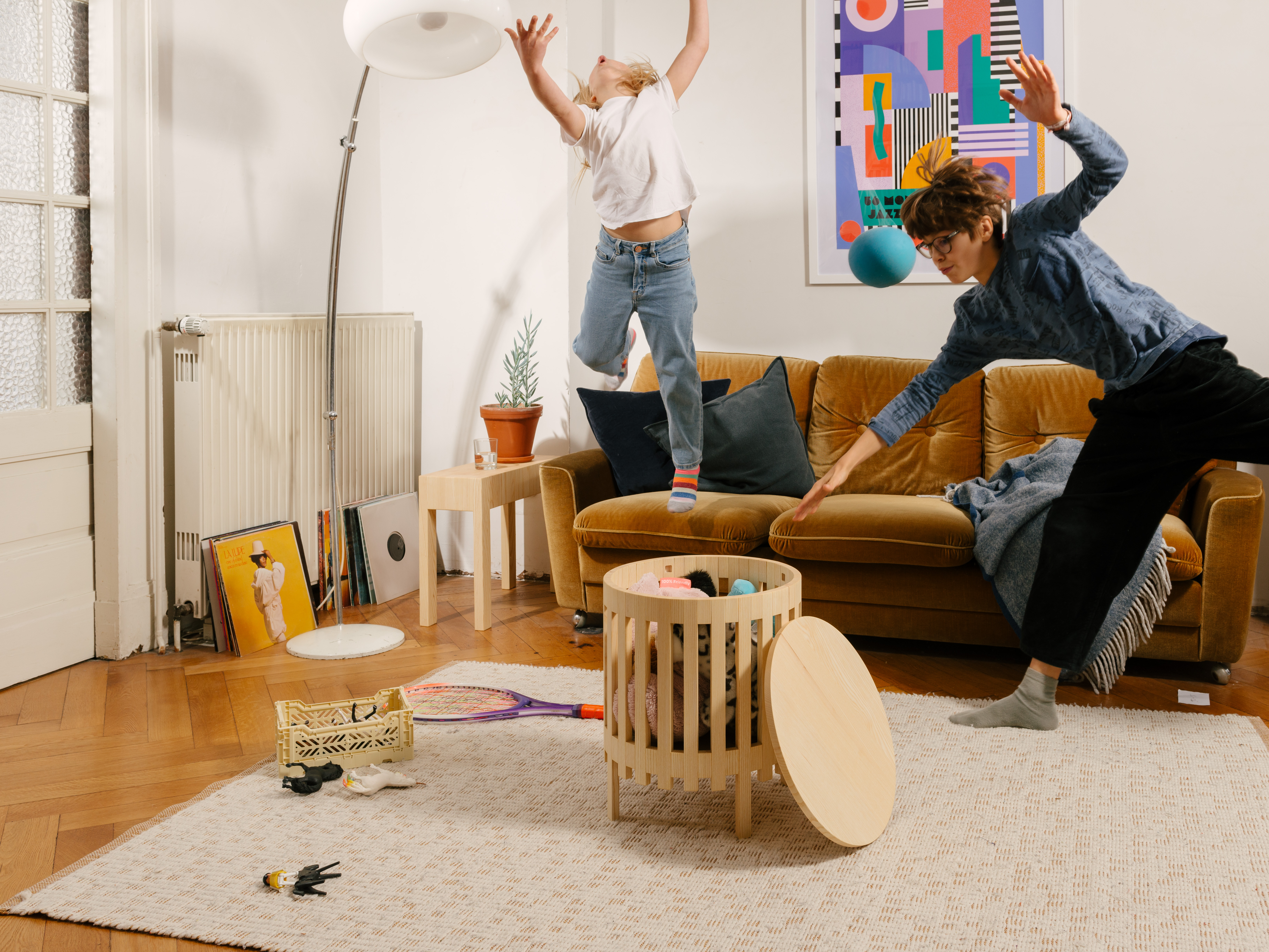 Ein modernes, gemütliches Altbau Wohnzimmer mit spielenden Kinder die in die Luft springen, in desssen Mitte ein wertiger Holzkorb aus Esche Vollholz mit Deckel als Couchtisch, gefüllt mit Stofftieren, steht.