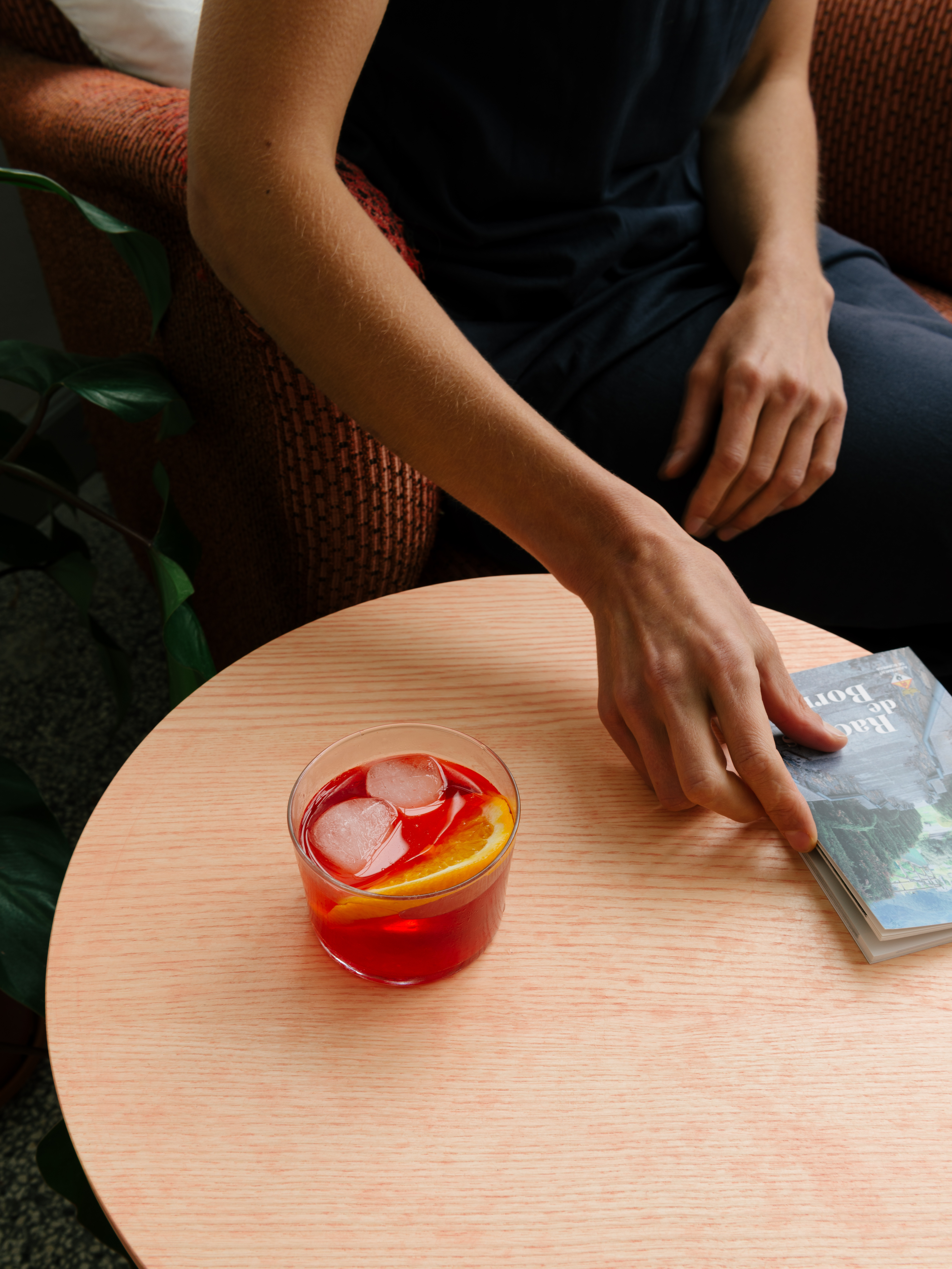 Ein Blick von oben auf einen runden Beistelltisch aus Esche Vollholz, erdbeerrosa lasiert, an dem eine Frau entspannt mit ihrem Buch und einem orangeroten Getränk sitzt.